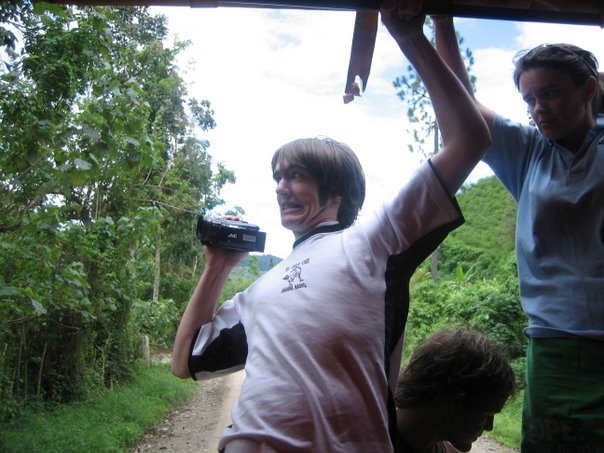 A young harry leans out the back of a truck, holding a small handycam as a concerned teacher looks on. Behind him we see part of a dirt road surrounded my lush greenery with a bright blue sky scattered with bright white clouds.