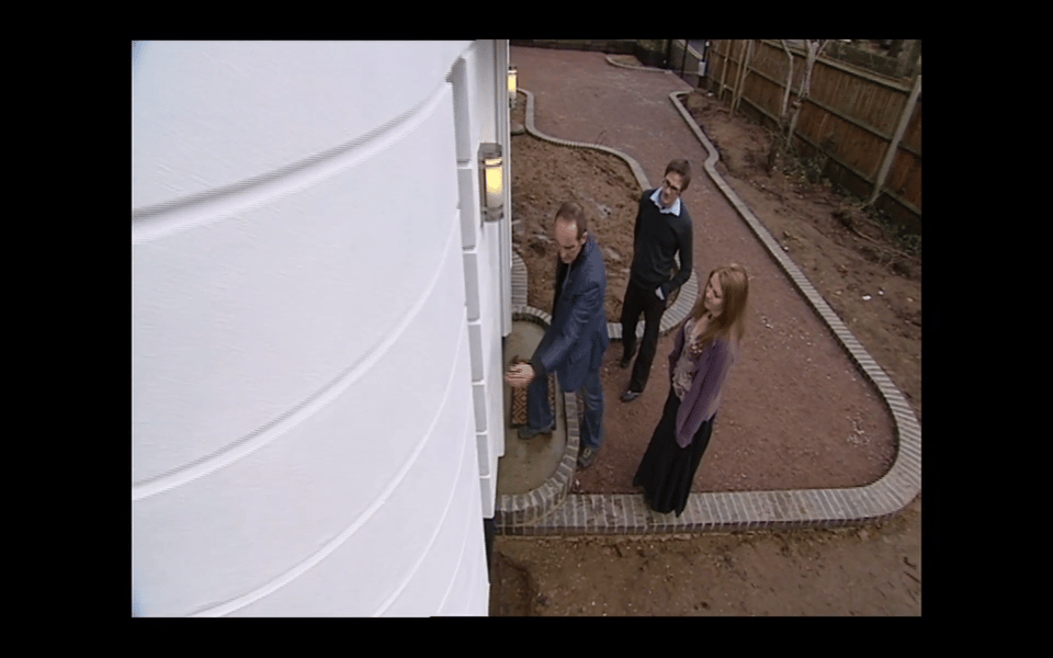 A screenshot of the show Grand Designs from 2007. A TV host touches a white wall as a couple watches