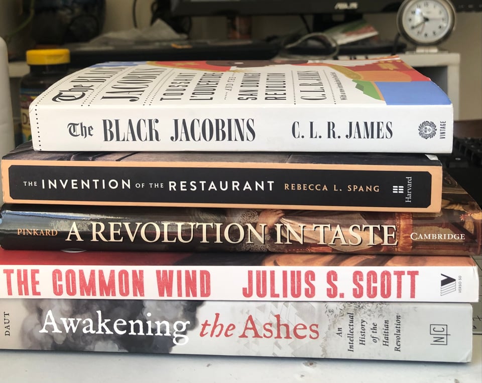 a stack of books: The Black Jacobins, by C.L.R. James; The Invention of the Restaurant, by Rebecca L. Spang; A Revolution in Taste, by Susan Pinkard; The Common Wind, by Julius S. Scott; Awakening the Ashes, by Marlene L. Daut