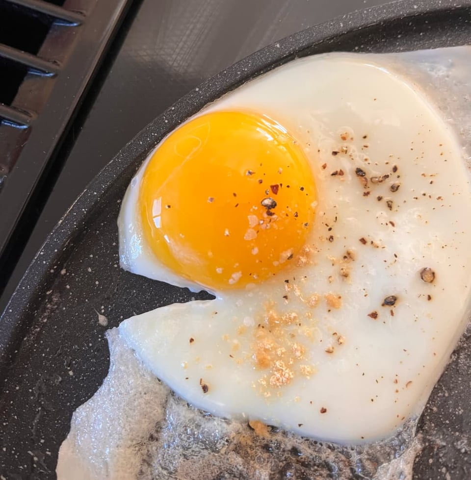 An egg frying on a pan. There is a small rip in the side of the egg directly under the yolk, and it makes the egg look like a fish with an open mouth.