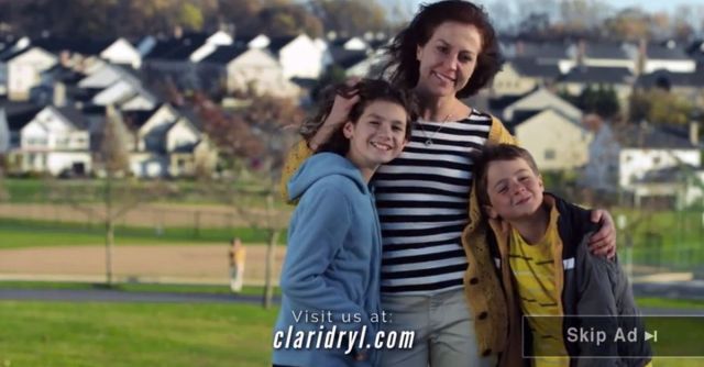 A woman stands outdoors with two children, smiling and posing for the camera against a backdrop of houses and a park.