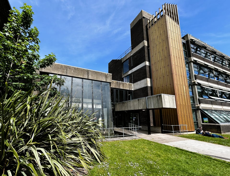 A complex Brutalist building. To the right is a four storey building with visible concrete floor plates and dark brick walls. On it's main elevation, the block switches to ribbon windows half hidden by brise soleil. A modern lift shaft has been added, clad in bronze metal. On the left, a two storey block with a concrete roof and glass walls creates the entrance. It includes a coverway over a level bridge that crosses a pool with fish in it (fish not pictured)