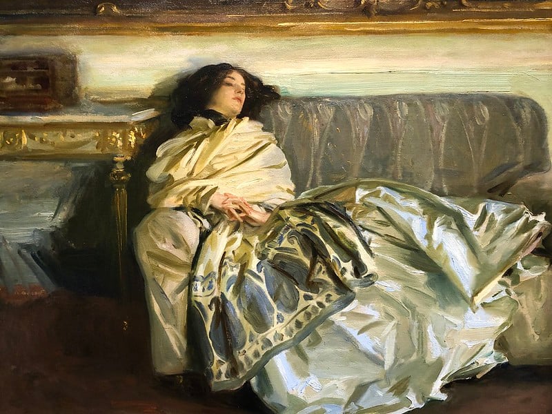 A painting by John Singer Sargent shows a woman with brown hair and wearing large skirts reclining on a couch looking tired