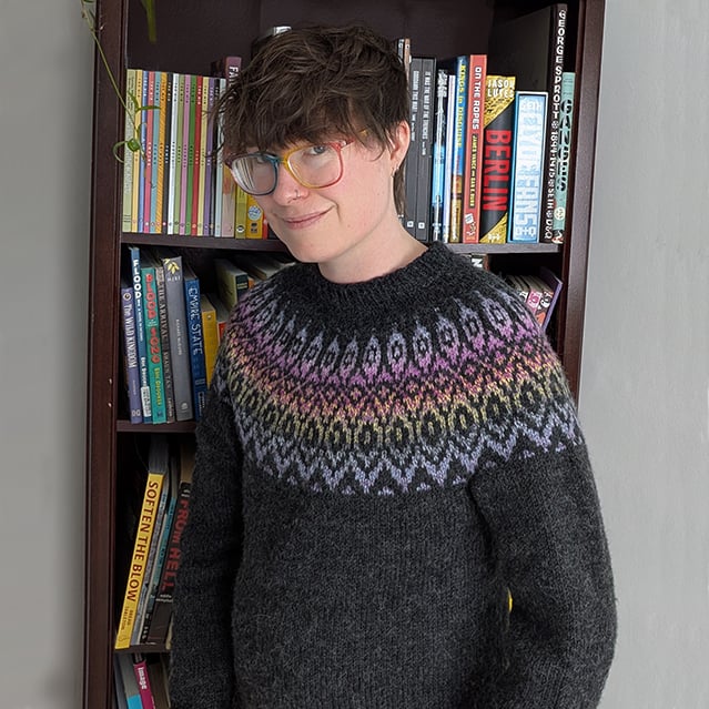 Maia stands in front of a bookshelf wearing a cozy charcoal knitted sweater with diamond patterned stranded colorwork across the chest and shoulders in a variegated yarn that shifts from pale blue to lavender to magenta to yellow and back to pale blue.