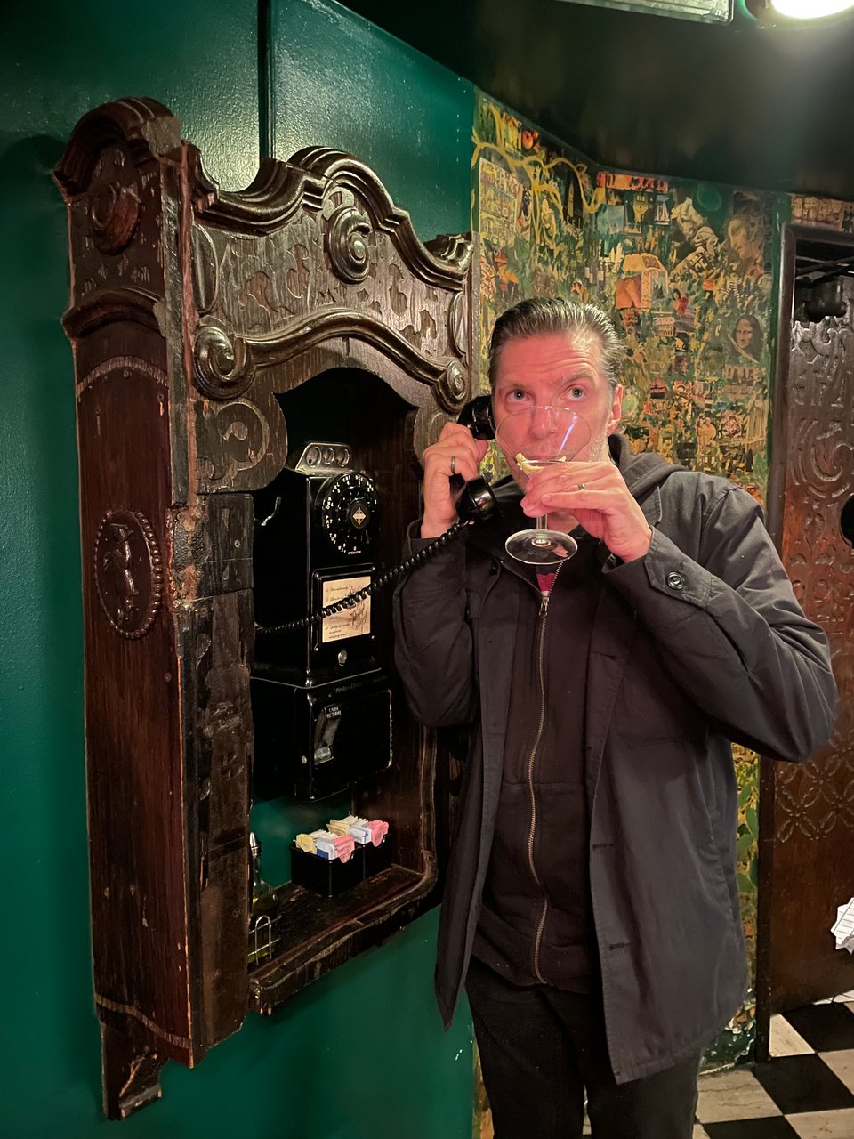 A man stands in a restaurant hallway holding an old payphone and sipping a martini