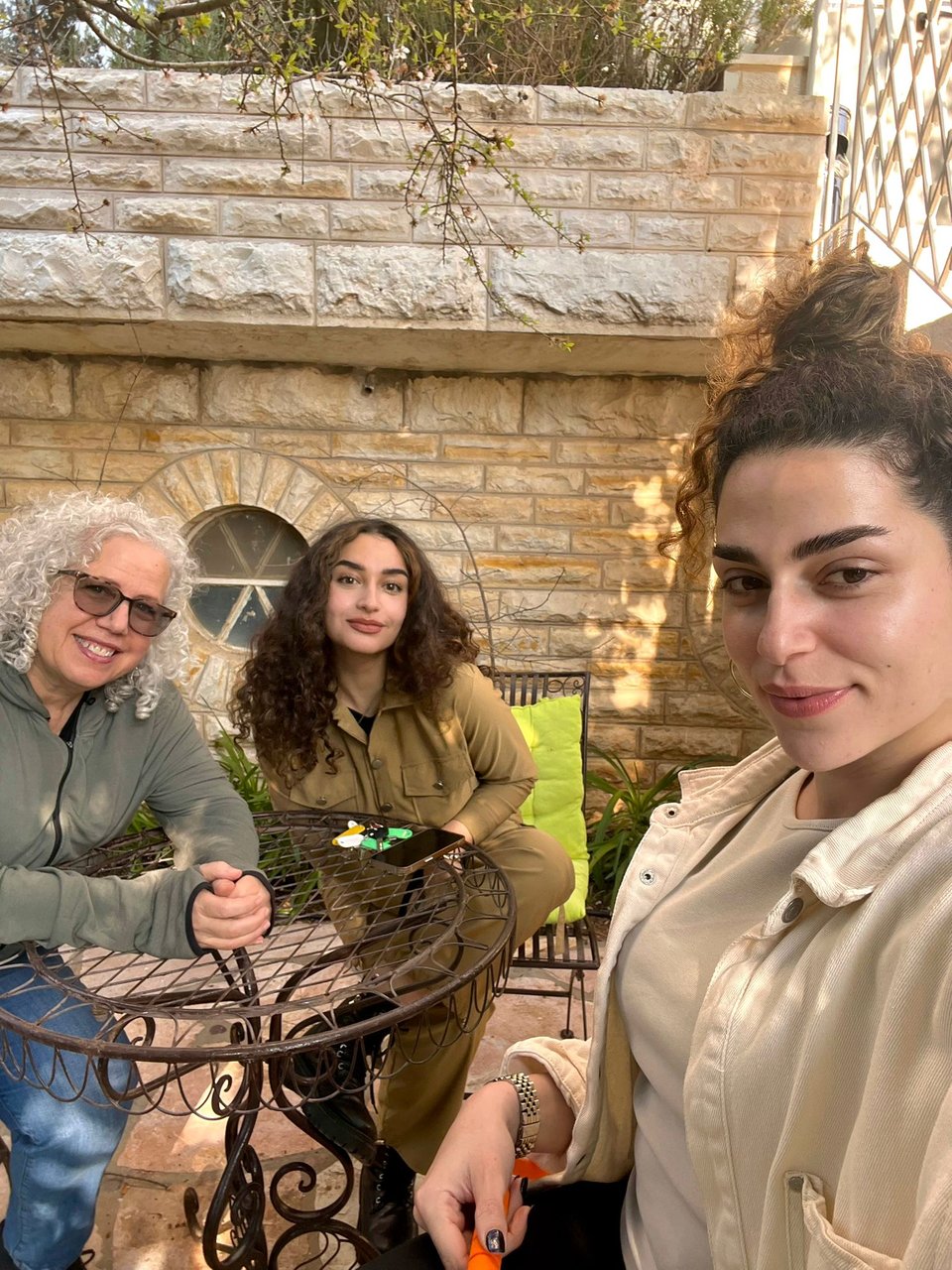 Three women sitting at an outdoor table in front of a stone wall
