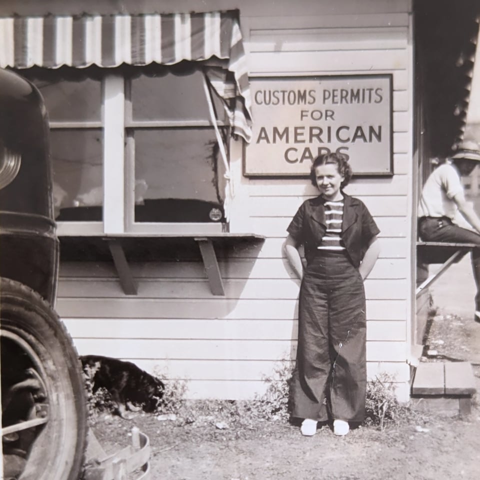 A young woman leans against a white wooden structure. She is wearing wide culottes and a matching blazer. She is beneath a sign that reads Custom Permits for American Cars. To the left, a dog sniffs the ground.