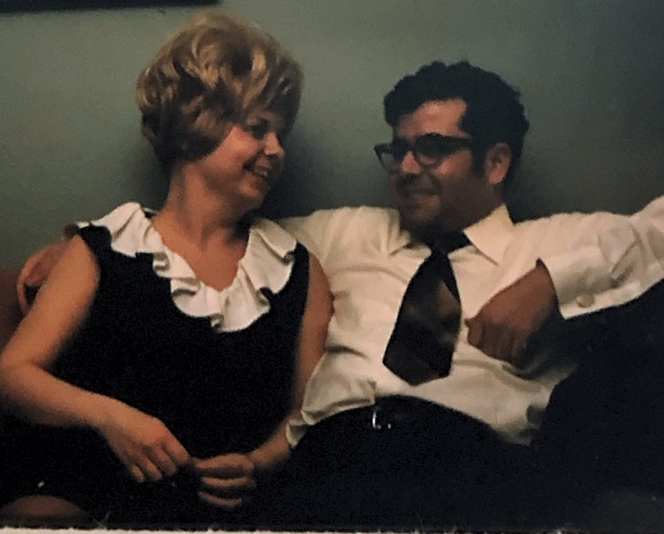 A white woman with a blond bouffant hair style is smiling and laughing and wearing a black dress with a white collar while looking at a man with black hair and glasses, who is also smiling and wearing a white button-down shirt and a tie. They are sitting on a sofa and looking at each other with happiness and relaxation.