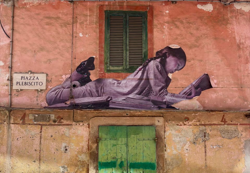 A weathered pink and ochre building facade features a large black-and-white paste-up mural of a child lying on their stomach reading a book surrounded by scattered papers, positioned beside a green louvered shutter above, with a white "Piazza Plebiscito" street sign mounted on the wall to the left and a green wooden door on the peeling lower level.