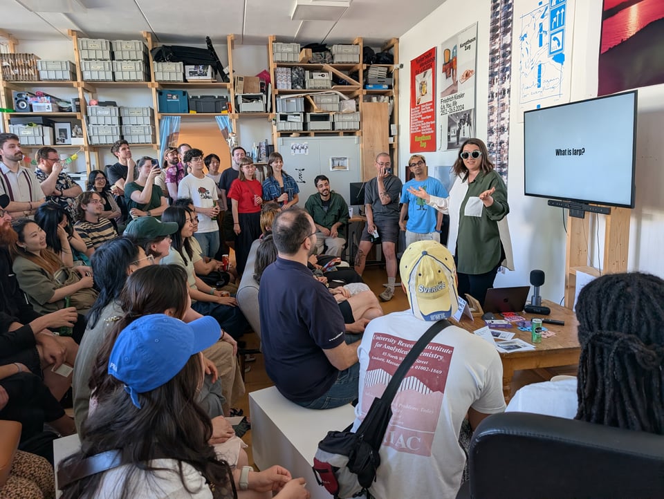 A woman in sunglasses lectures to an amused crowd. The TV screen next to her says "what is larp?"