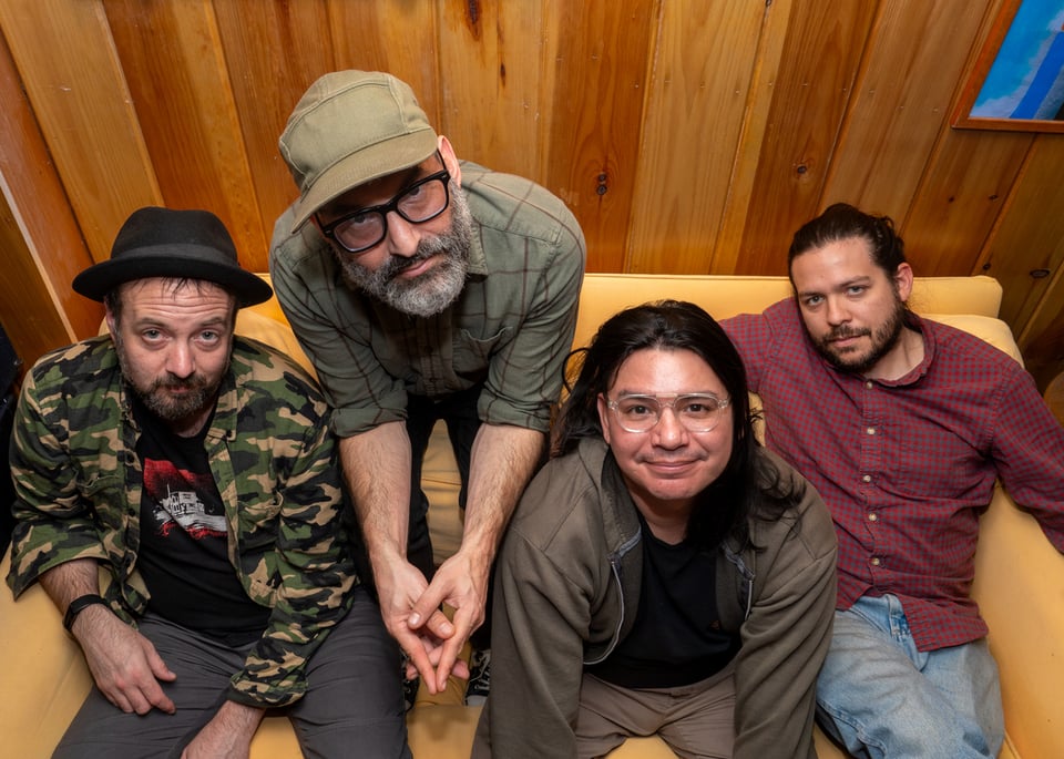 Four men sig on a yellow couch in front of a wood-paneled wall, looking up at the camera.