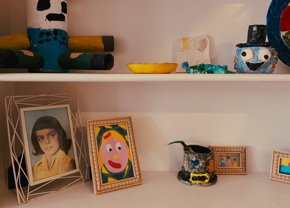 Shelf displaying colorful handmade crafts and paintings. Includes a vintage child's portrait, abstract art, a smiling face, and painted clay figures.