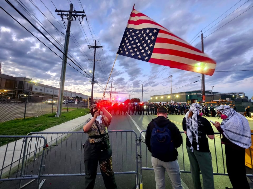 A protestor flies an upside down US flag across from a riot line of police officers near Delaney Hall. The sky is a light blue as the sun, sets with a scattering of clouds. Behind the police line in the distance a number of emergency vehicle lights backlight the line of officers.