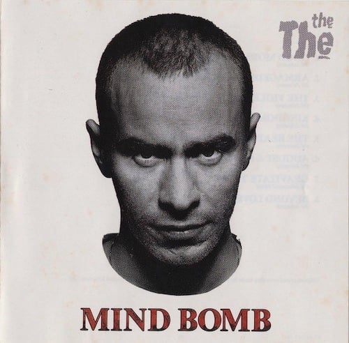 A headshot of The The's Matt Johnson, a white man with his head shaved short, with a receding hairline. He has a dark brow and pointy stubble. He has a very serious expression on his face, but with a slight wryness, which if you've heard his music, makes sense. The photo has had the background removed, so his head appears to float in a white blank nothing.
