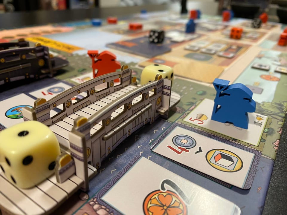 White dice on either end of a small white carboard bridge.  Cards and blue and red meeples are in the background.