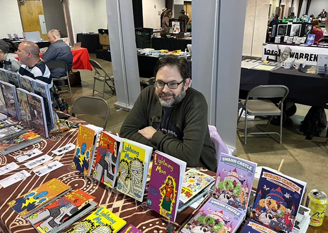 A middle-aged cartoonist sitting behind a table that is covered with comics for sale.
