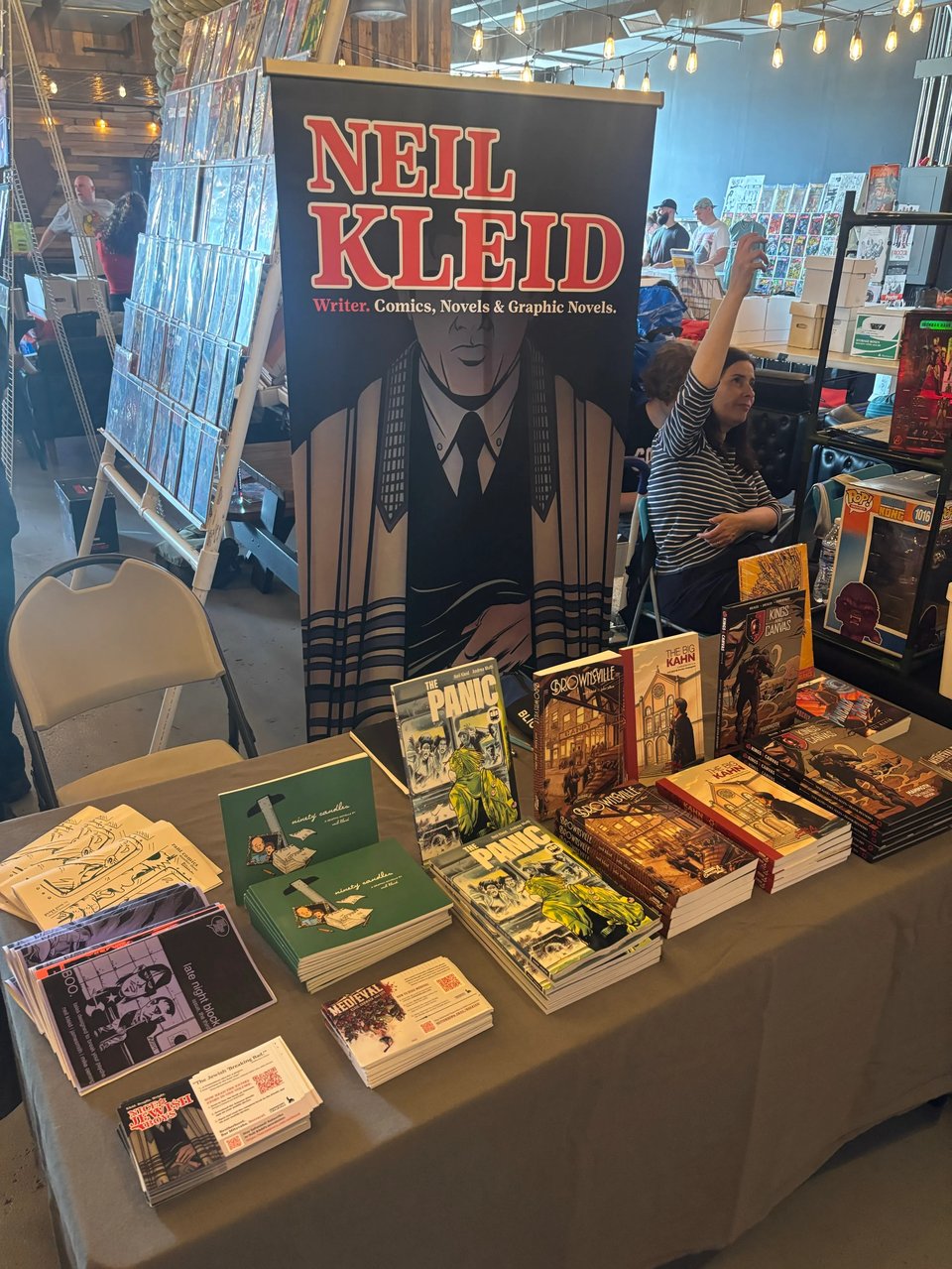 neil's table setup at staten island comics fest, featuring a banner with his name on it and an image from nice jewish boys, and a bunch of assorted comics and cards