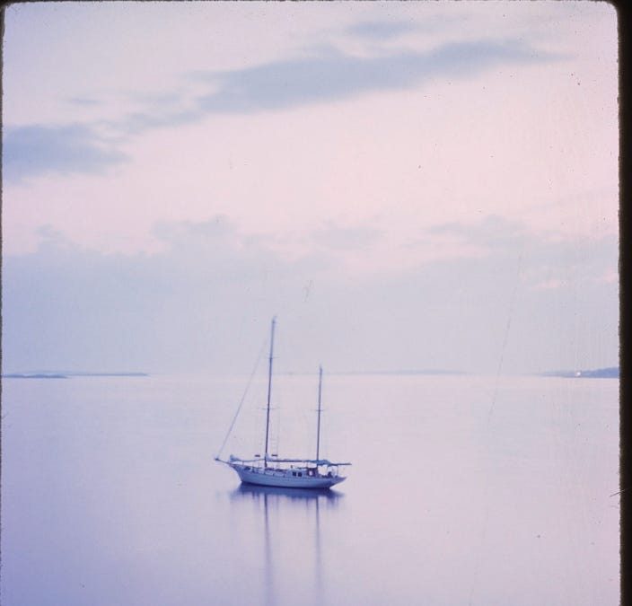 A single sailboat resting on calm, pale lilac water under a hazy pink sky, on a faded vintage photograph