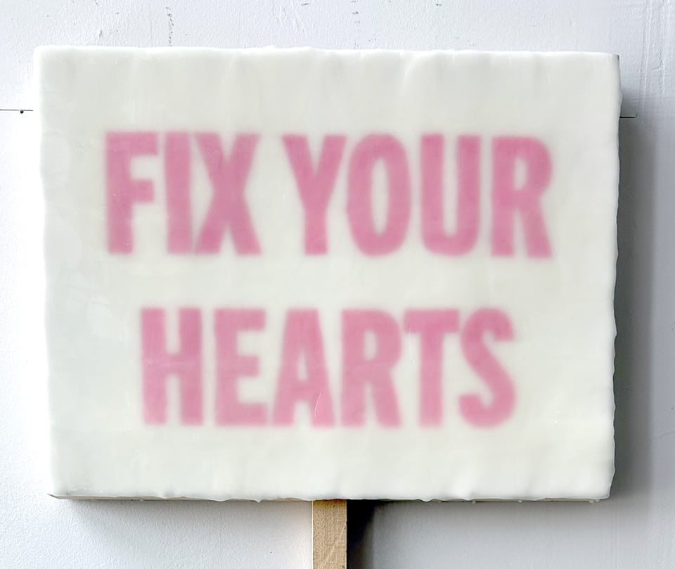 Little painting. White background. Pink letters that say FIX YOUR HEARTS. There's a stick at the bottom so it looks like a protest sign.