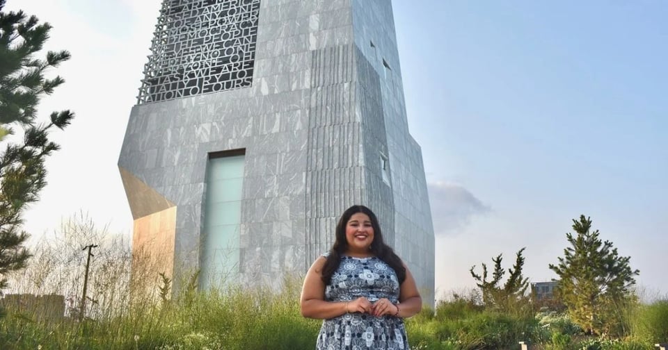 A woman with long dark hair stands smiling in front of a tall, modern monument made of gray stone panels. The structure features engraved text and geometric design elements, surrounded by greenery under a clear sky.