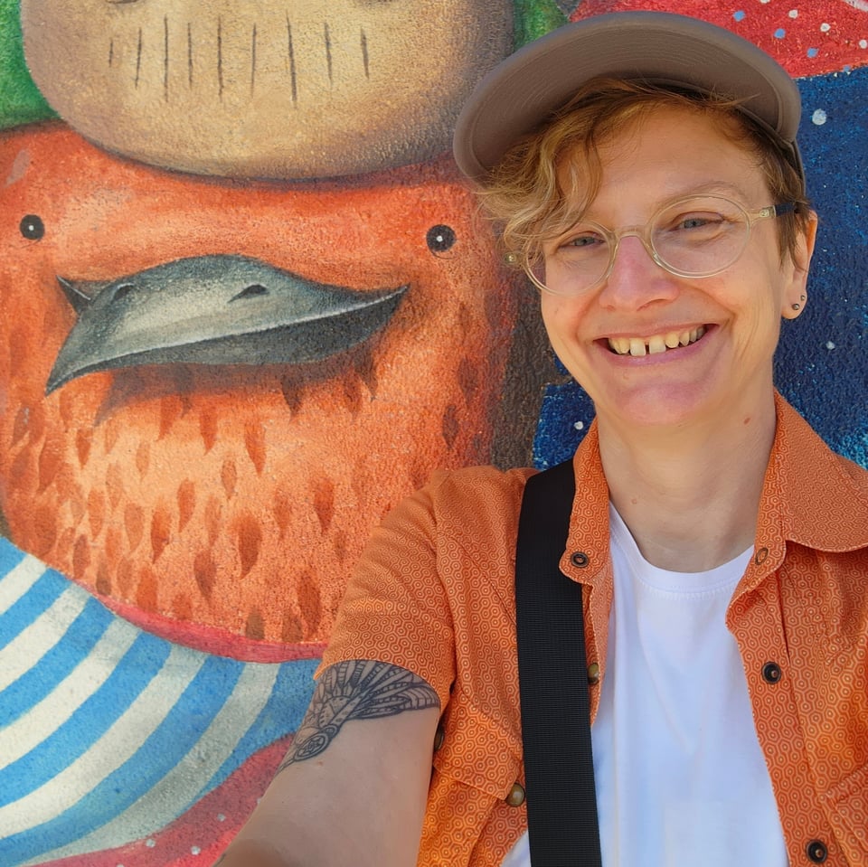 It's me, a 52-year old white nonbinary person with a forelock of blonde hair and glasses and a goofy smile. I'm standing next to the mural of a smiling bird of some kind. We look like really good friends.