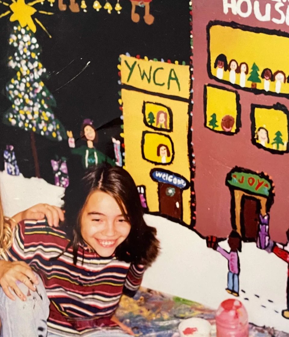 Photo of a young Puerto Rican girl, smiling with paint on her hands and shirt, in front of a hand painted mural.