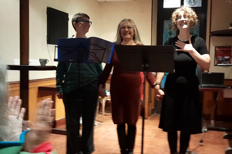 Dans une salle de bar. Trois personnes de face qui ont un grand sourire. Devant elles, des pupitres. Elles se tiennent les mains. Celle de gauche regarde celle de droite. Celle du milieu regarde devant elle. Celle de droite regarde devant en se tenant une main sur la poitrine. En bas à gauche de l'image, on voit des mains en train d'applaudir.