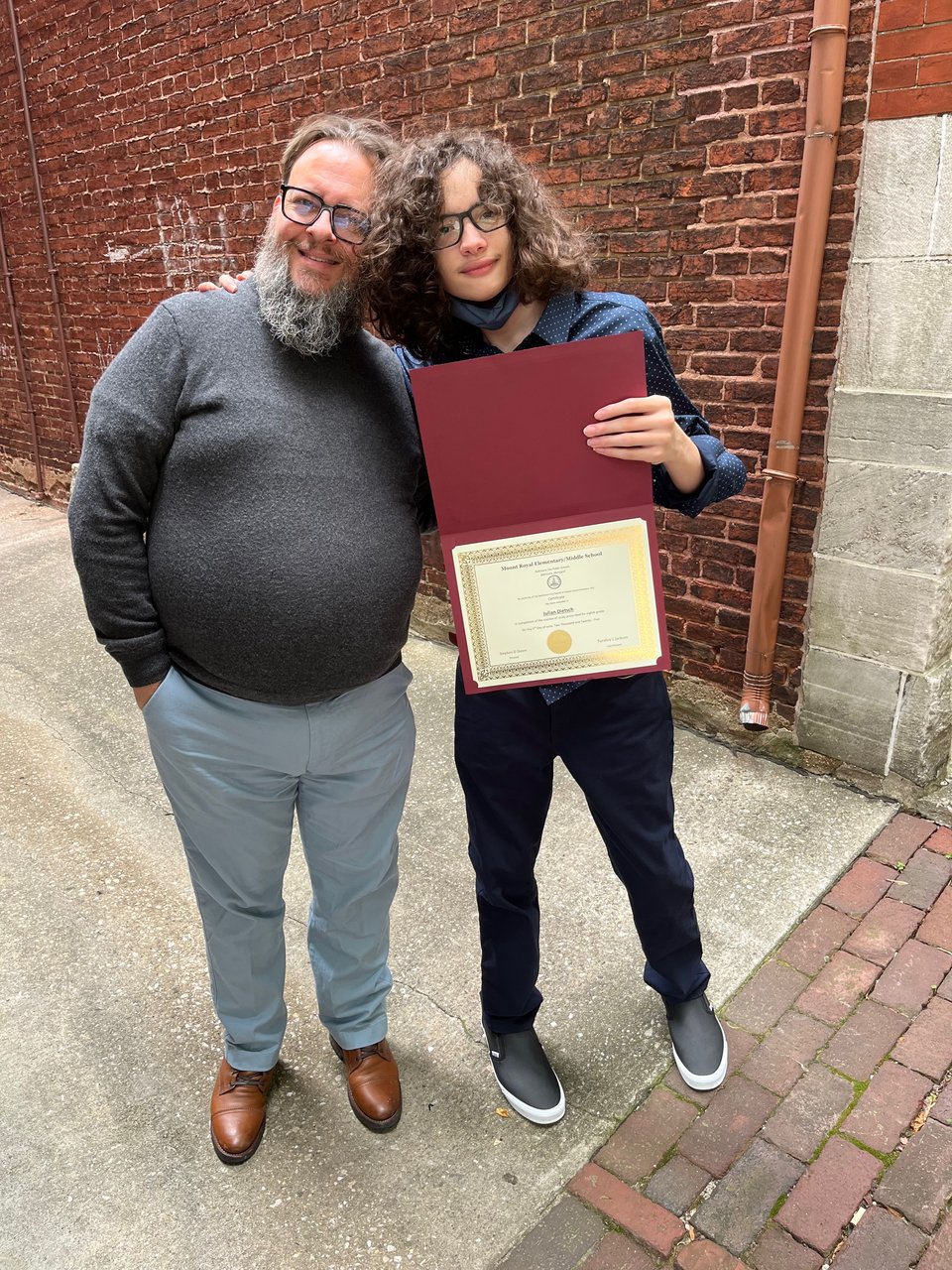 a man stands with teenaged boy, holding a graduation certificate