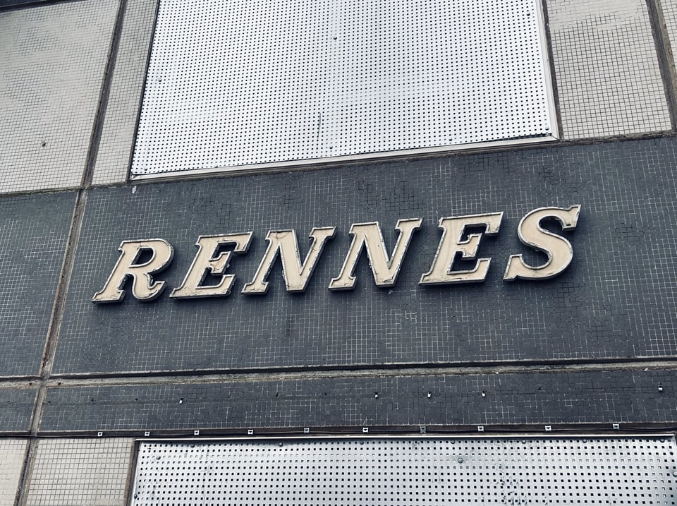 Close up on the large 1960s letters that spell out RENNES over the door of the block. The doorway below and window above have been boarded up with sheet metal.