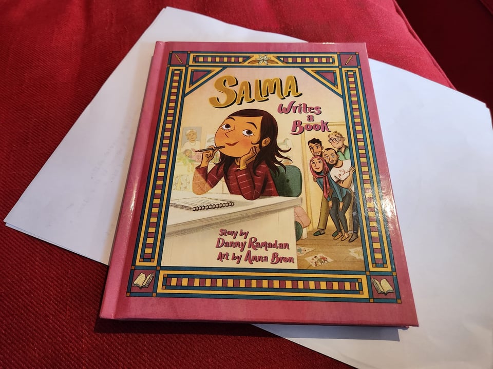 A pink book with an illustration of a young Syrian-Canadian girl at a writing desk, called, Salma Writes a Book, by Danny Ramadan, art by Anna Bron