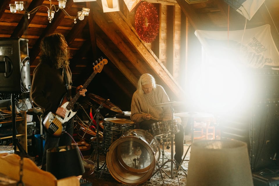 A man with long brown hair and a beard plays a bass guitar to the right of a man with chin-length white hair sitting at a drum kit.