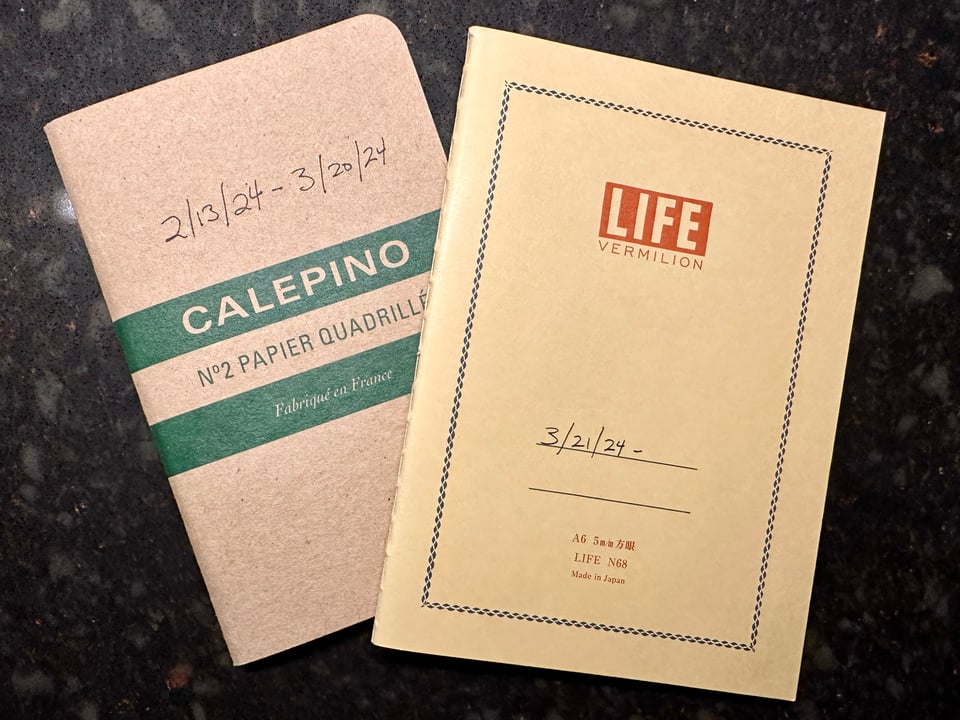 Photo of 2 notepads on a black faux-marble surface. One notepad is dated 2/13/24-3/20/24, and the other is dated 3/21/24-