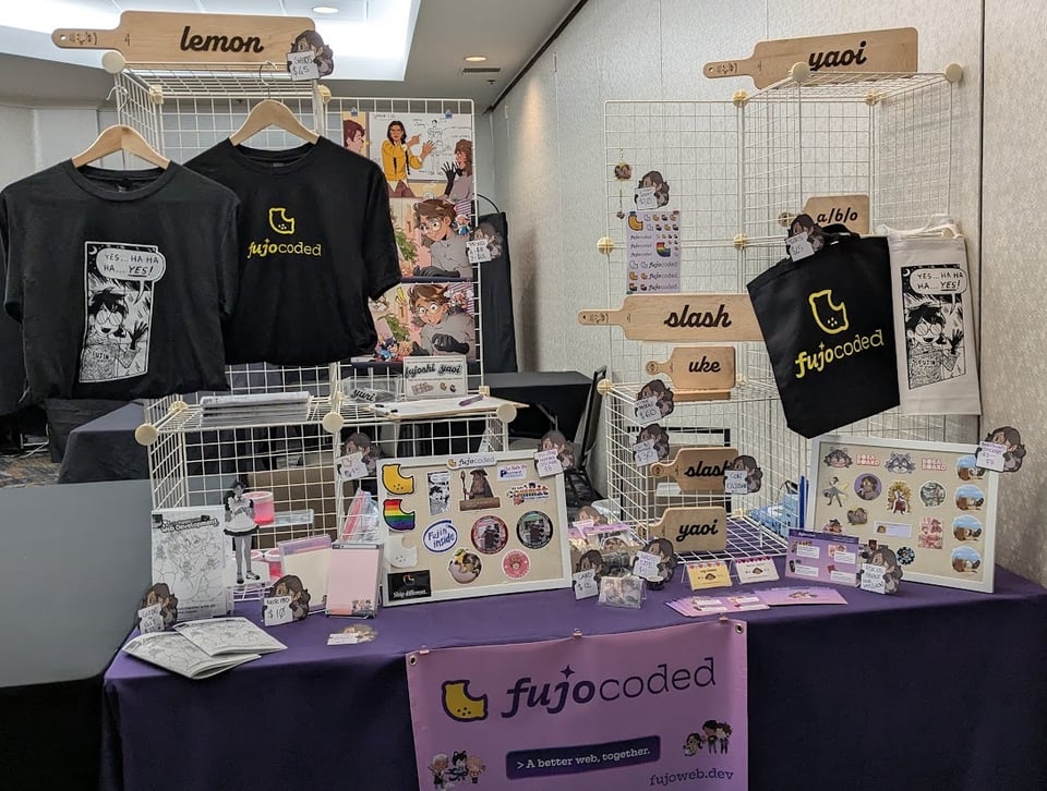 Photo of a dealer's table in a hotel meeting room. A small professionally printed banner with the FujoCoded logo and the slogan "A better web, together" hangs on the front of the table, which is covered in a purple tablecloth. Cream-colored wire cubes in 3 by 2 stacks are arranged on either end of the table to create surfaces for merchandise. On the left cube stack are Fujin Sickos t shirt, a FujoCoded lemon logo shirt, 3 FujoCoded art prints, a newsletter signup sheet offering free stickers in exchange for signup, several containers of catboy bathwater, and a hime-size cheese paddle that says 'lemon'. On the right cube stack are several hime and chibi size cheese paddles, a Fujocoded logo tote bag and a Fujin sickos tote bag, some phone charms, and a page of FujoCoded logo stickers. In front of the left cube is an array of printed-out FujoGuides with a standee of HTML as a paper doll wearing a maid outfit. In the middle front of the table is a small bulletin board offering an array of FujoCoded stickers from the FujoCoded kickstarter. On the right side of the table is an array of business cards and info cards about FujoCoded's different projects, followed by another small bulletin board of older BobaBoard stickers.