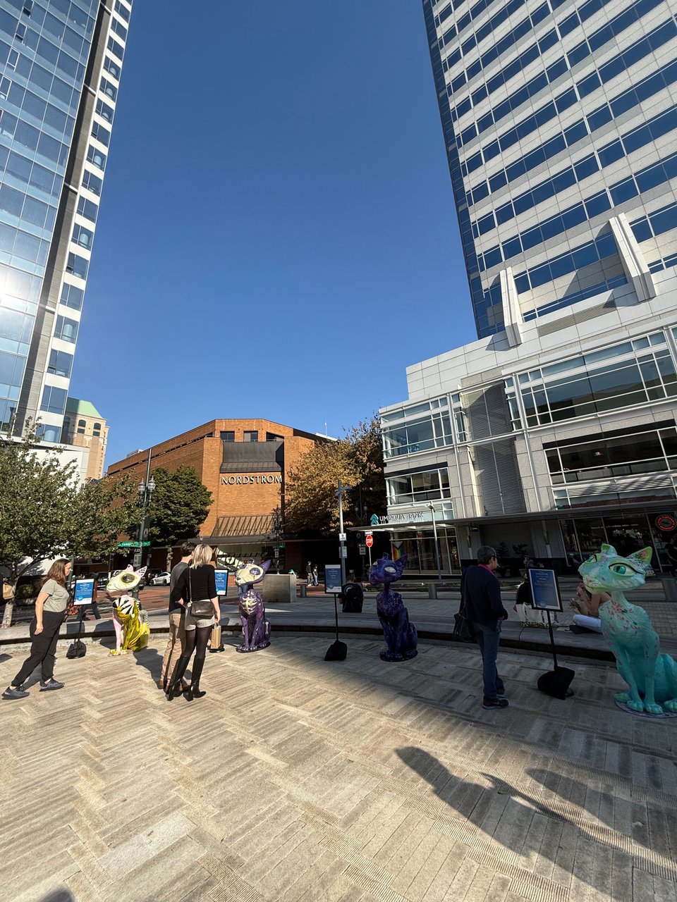 People walk amongst and take photos of large cat sculptures, surrounded by tall buildings.