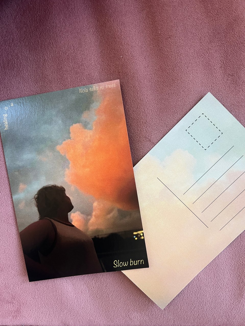 Front and back of Slow burn postcard against rose velvet. The front is a picture of Nola in shadow looking up at vivid rose gold clouds in a grey-blue sky with "Slow burn" written in yellow letters in the lower right corner. The back shows a transparent close-up of the clouds and sky.
