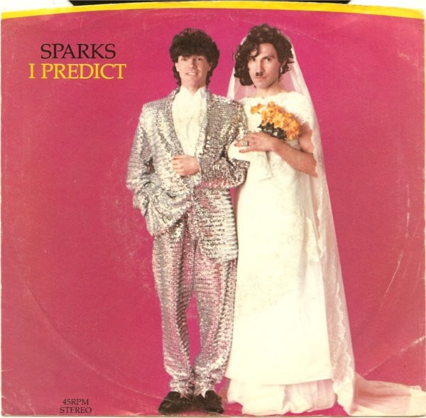 The cover for the 45 of Sparks’ single “I Predict.” It shows Russell Mael, a white man with big curly brown hair, in a sparkling sequined(?) suit. He is arm and arm with a bride in full bridal gown, played by his brother Ron Mael, who looks a lot like his brother but has his famous Hitler-esque mustache. Ron holds a bouquet of yellow flowers.