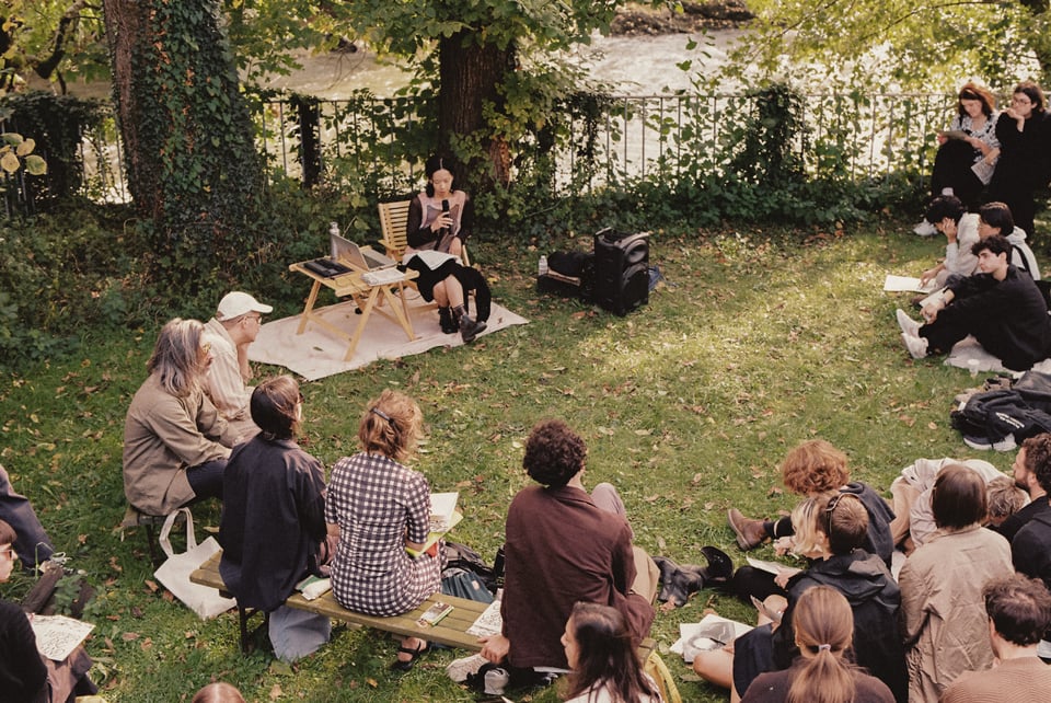  [A semi-circle of people sit on a grassy outdoor space before a river. Charmaine reads into a microphone at the front, shaded by a large tree.]