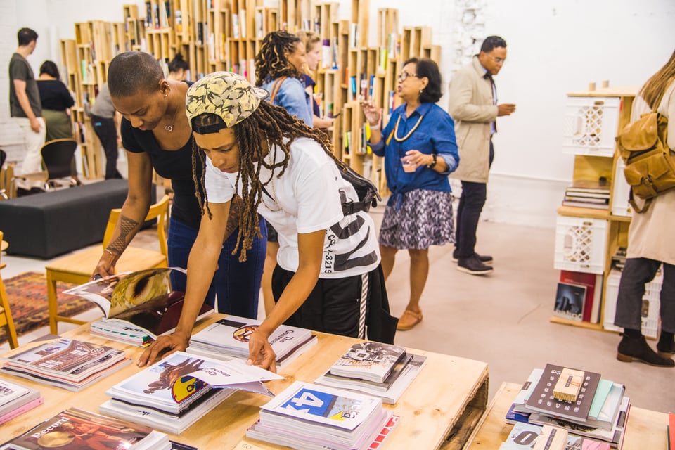 Two people look through books displayed on a table, while others mill around behind them. Slender wooden boxes stacked on top of each other at different heights make up the bookshelves in the space, giving them the look of organ pipes.