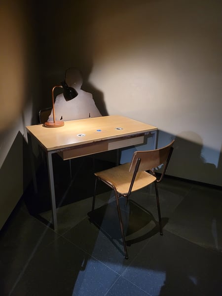 A wood chair sits in front of a wood desk with a lamp on it. Behind the chair is a silhouette of an interrogator.