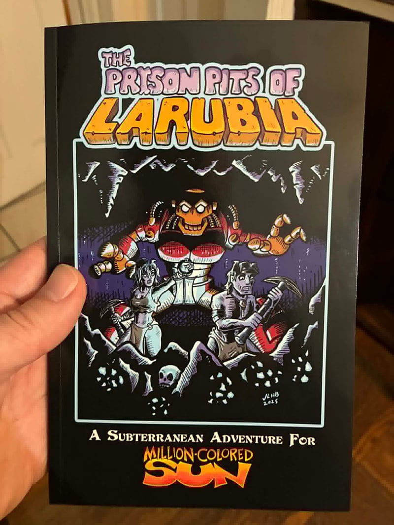 My hand, holding up the printed copy of Prison Pits of Larubia, with luscious art and cover design by Josh Burnett.