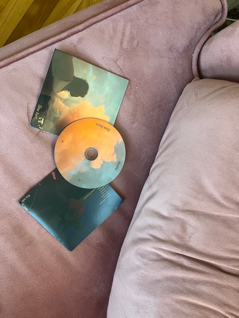 Slow burn CD with sleeves showing the front and back album art. The CD has rose gold clouds on a dusty blue sky with the words "healing is a Slow burn" written in dark gold. The front cover shows a picture of Nola in shadow looking up at the clouds with "Slow burn" written in small yellow letters in the lower right corner. The back cover shows a dark picture of Nola holding a guitar in deep shadow and dim firelight with the track list and "Nola talks to trees" written in small yellow letters. The background is a rose velvet loveseat.