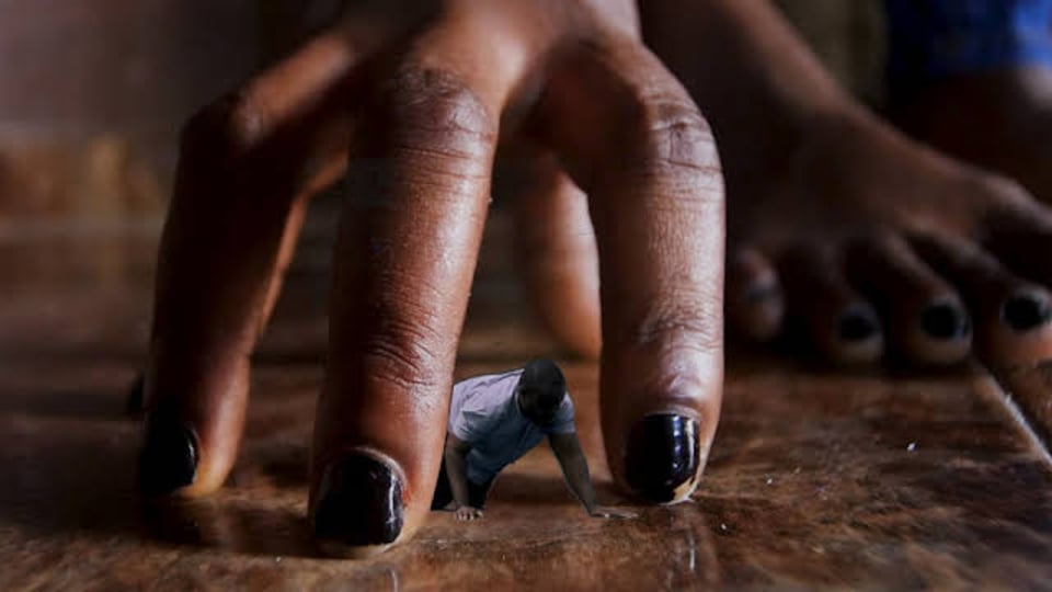 A shrunken Black man crawls on a polished wood floor, within the grasp of a Black woman's huge fingers spread around him like a cage.
