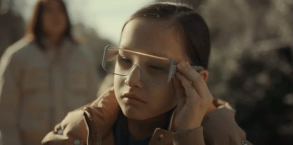 A child wearing futuristic-looking surveillance glasses puts her left hand up to the glasses, perhaps to activate a feature.