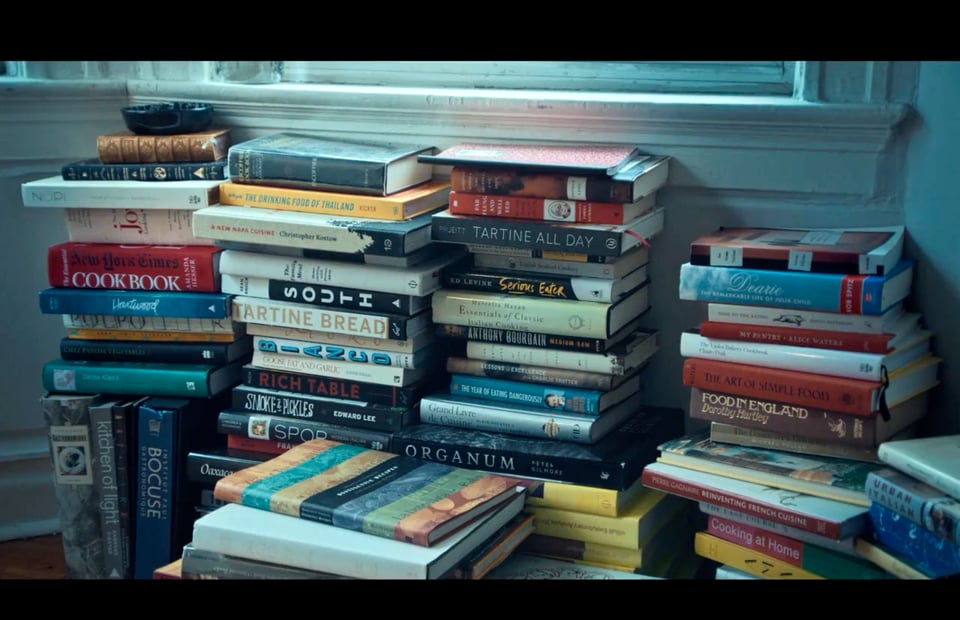 A number of cooking related books stacked in piles on the floor of an aparment.