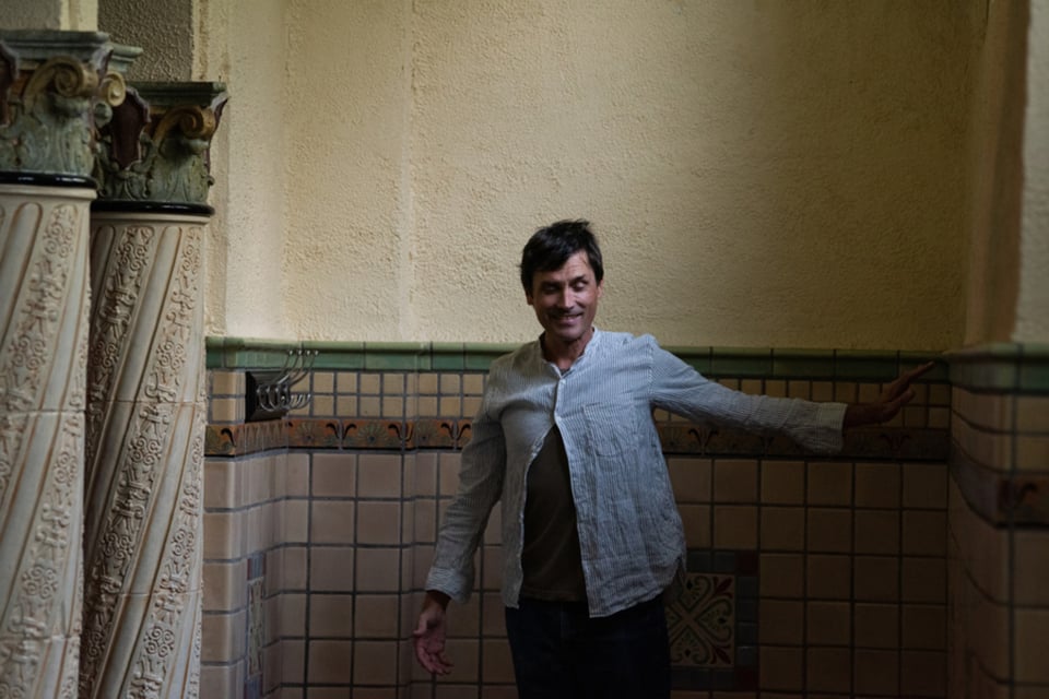 A man stands in a tiled alcove with his left arm extended to the side, with a pair of columns to his right.