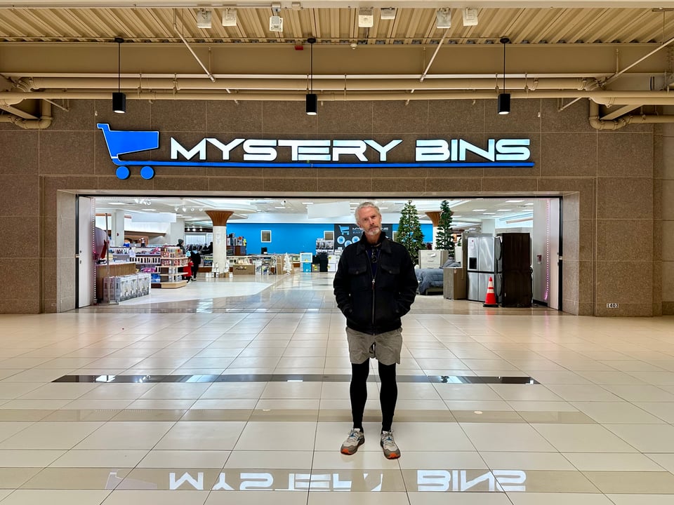 Photo of a white man standing in front of a mall store called MYSTERY BINS with a puzzled expression on his face
