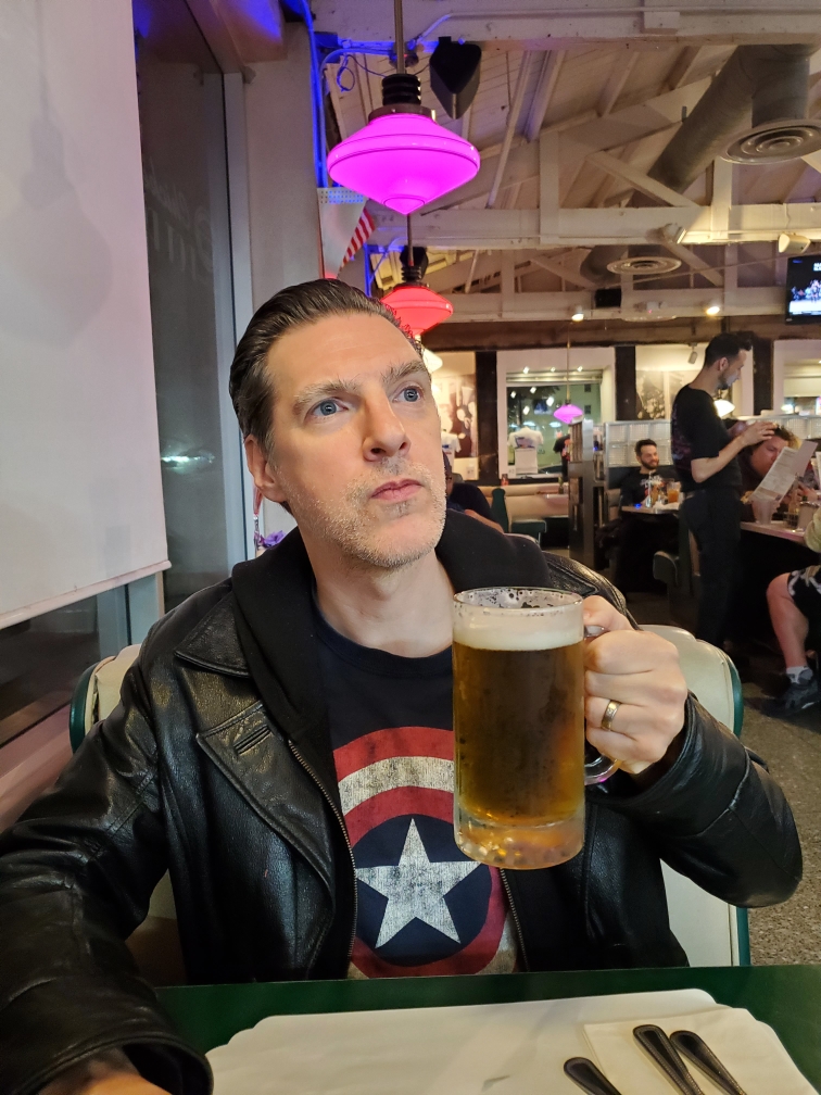 A man in a Captain America shirt holds a large glass of beer in a diner and stares up in inspiration
