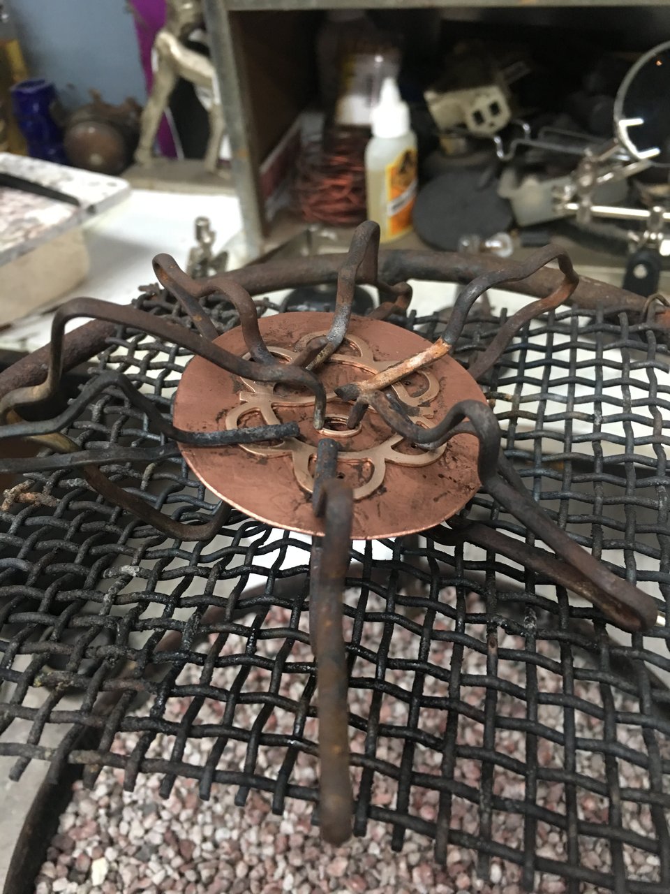 A hand cut brass circle pendant with a separate Taino design is clasped together over a grate in the process of soldering