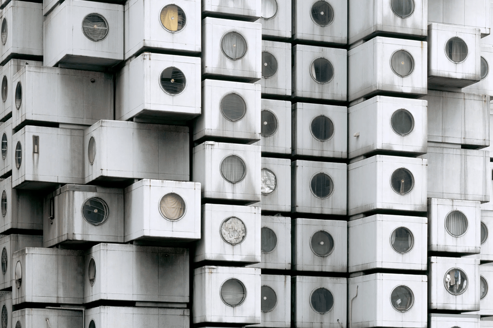 A close-up of a building that is composed of light gray units, each one with a dark window. Some of the windows have glass in them, while others appear to have been broken in the past, and are now covered up with fabric.