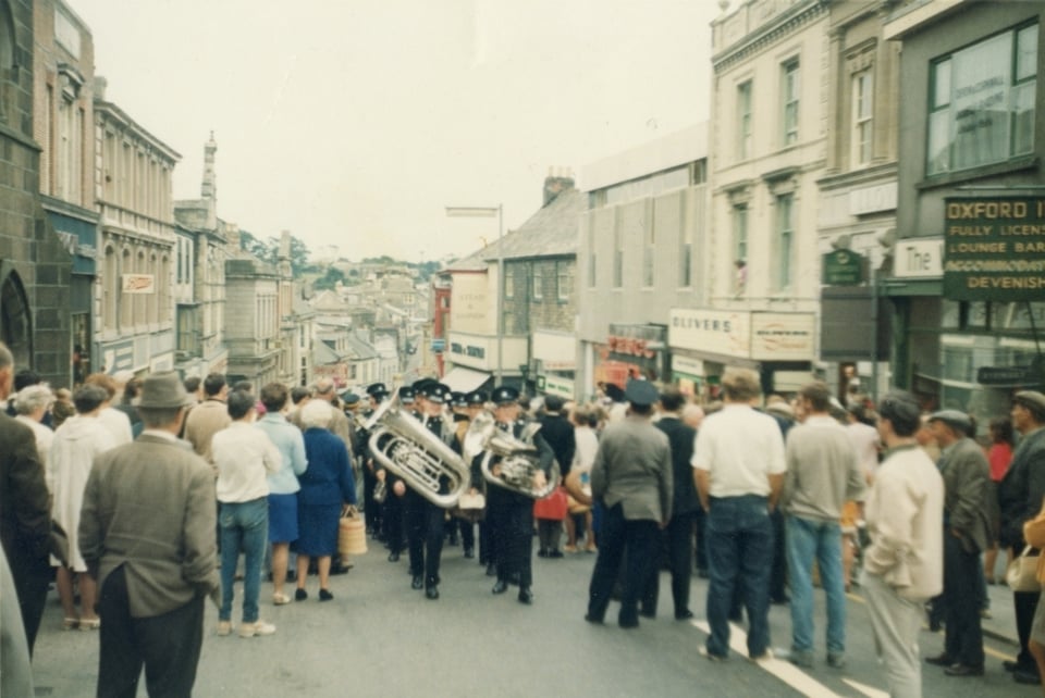 A faded photo of a brass band walking up a slope, through a crowd of people. All the people are looking down the slope, where the top of the Tesco building is visible.